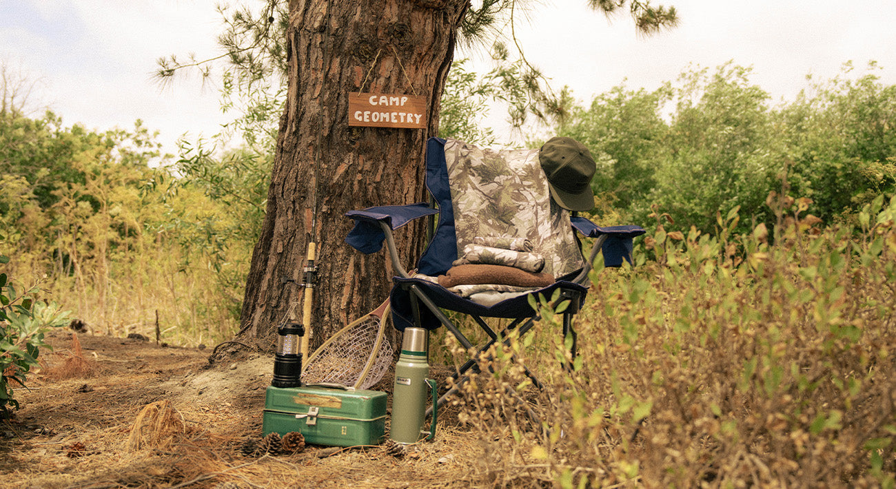 A folding chair with a hat, towels, and fishing net sits by a tree with a Camp Geometry sign. Nearby are a green lunchbox, thermos, water bottle, and dried grass and bushes.