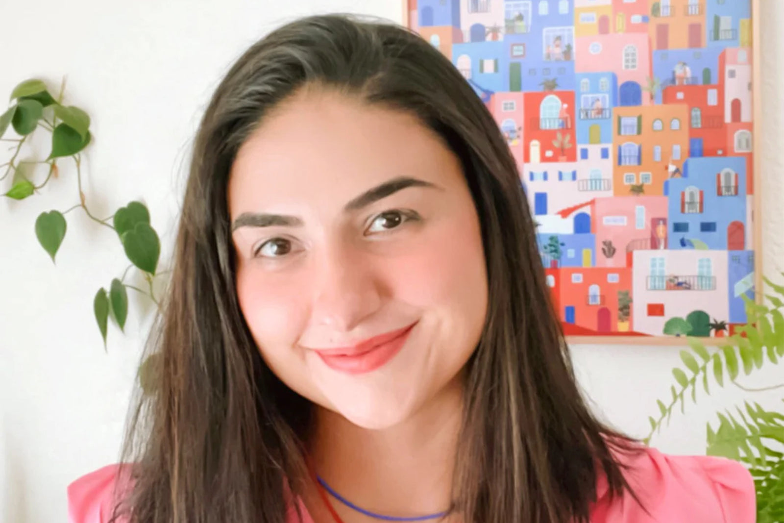 A woman with long dark hair and a pink top smiles at the camera. Behind her is a colorful painting of buildings and some green plants on the wall.