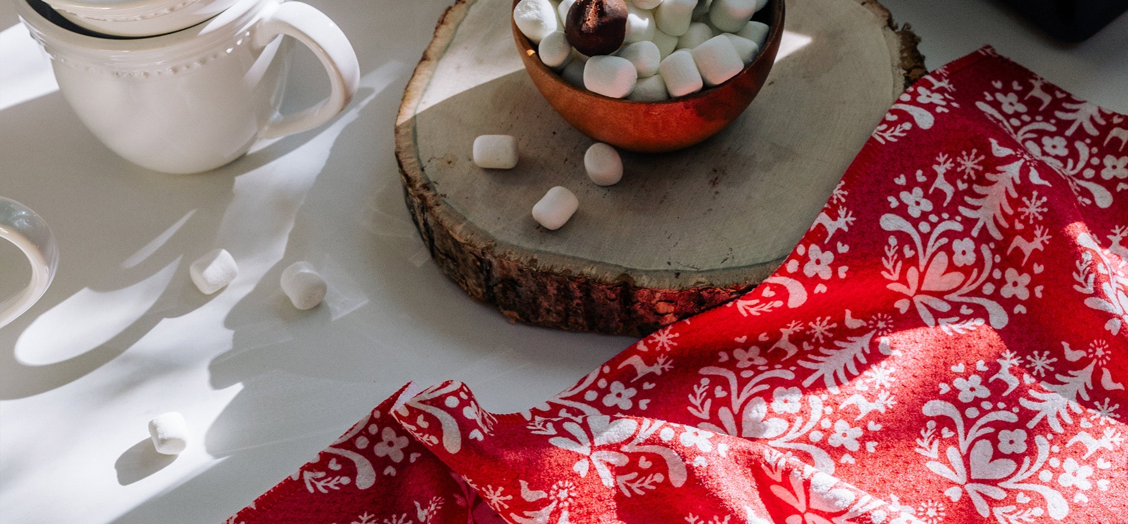 A wooden slab holds a bowl of marshmallows and cocoa beans. Scattered marshmallows and a white mug sit nearby on a white surface with a red and white patterned cloth.