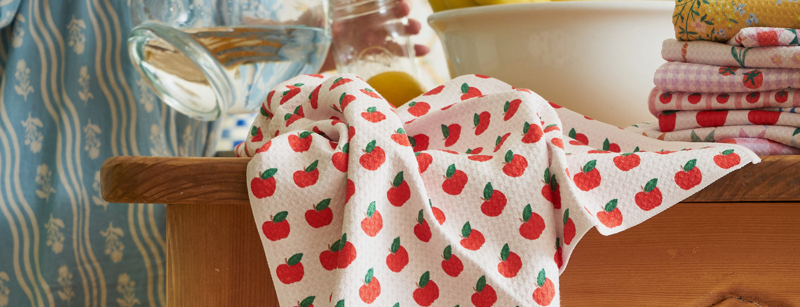 A white dish towel with a red apple pattern drapes over a wooden counter beside a white bowl, a water pitcher, and a stack of folded towels in a cozy kitchen setting.