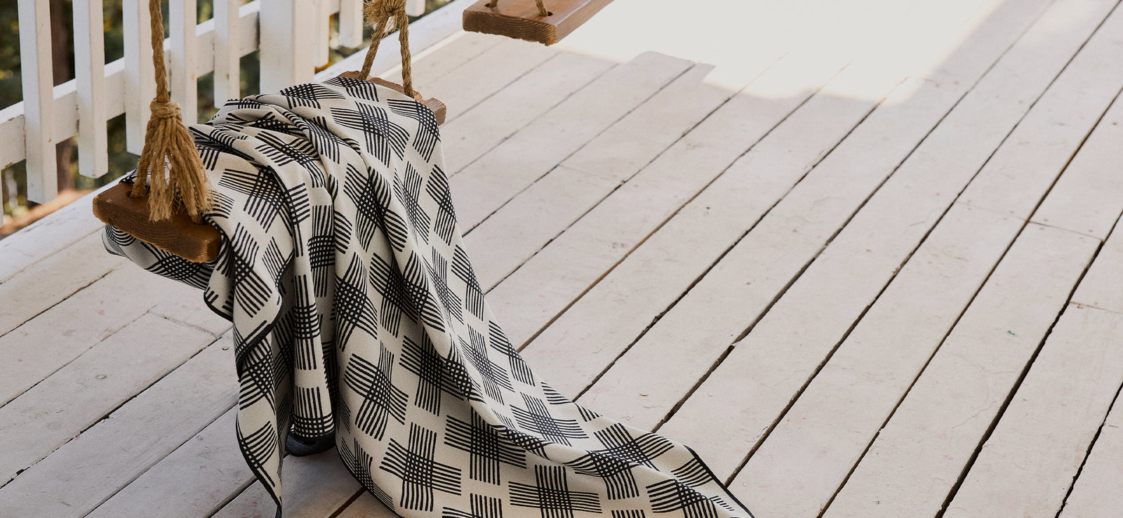 A black and white patterned blanket draped over a wooden swing with thick rope on a white-painted wooden porch, next to a white railing. Sunlight casts soft shadows across the scene.