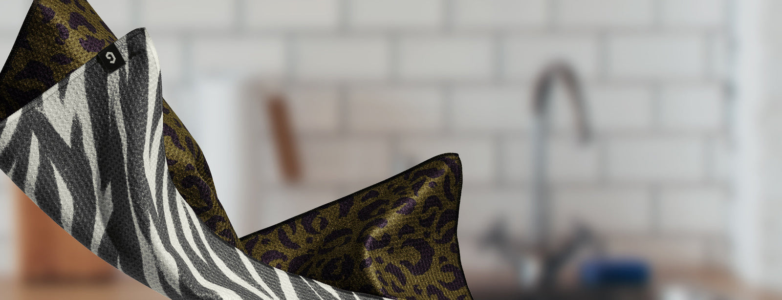 Close-up of folded fabric with zebra and leopard print patterns; a small black tag is visible. The background is a blurred kitchen scene with white tile and a faucet.