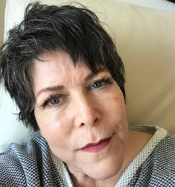 A woman with short, dark, wavy hair and green eyes is looking at the camera, wearing a gray and white knit top and light makeup, sitting against a cream-colored background.