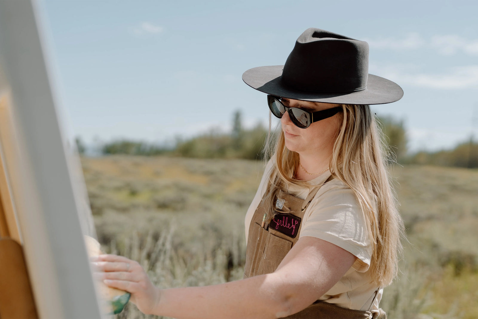 A woman wearing a wide-brimmed hat, sunglasses, and overalls paints on a canvas outdoors in a field, with greenery and blue sky in the background.