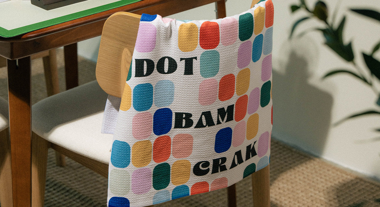 A colorful towel with oval patterns and the words DOT BAM CRAK hangs over a wooden chair next to a table. Green plant leaves are visible in the background.