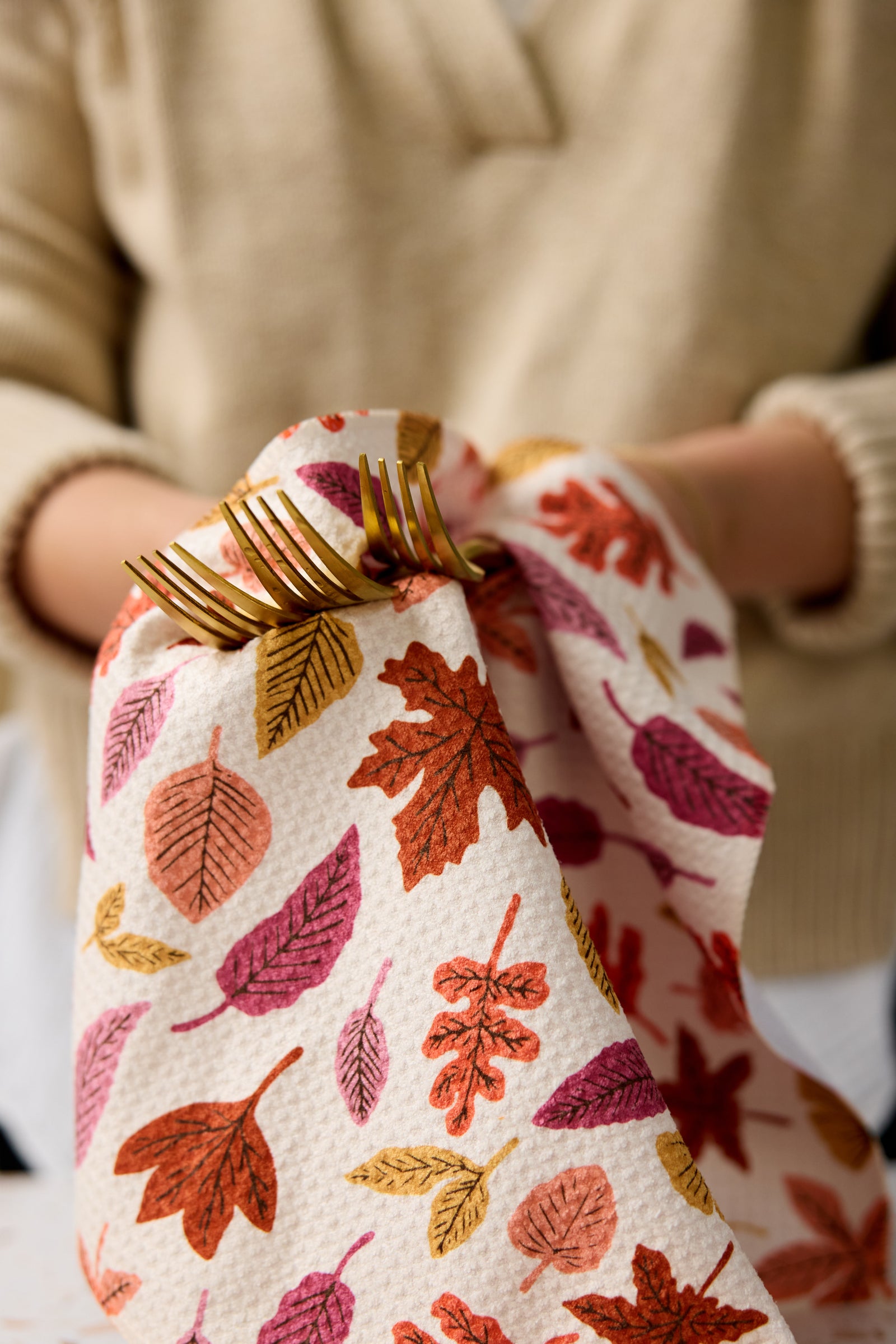 A person holding gold forks wrapped in a napkin with a colorful autumn leaf pattern; only their hands and beige sweater are visible.