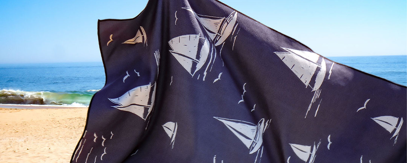 A person holds up a navy blue beach towel with white sailboat designs on a sandy beach, with waves and a clear blue sky in the background.