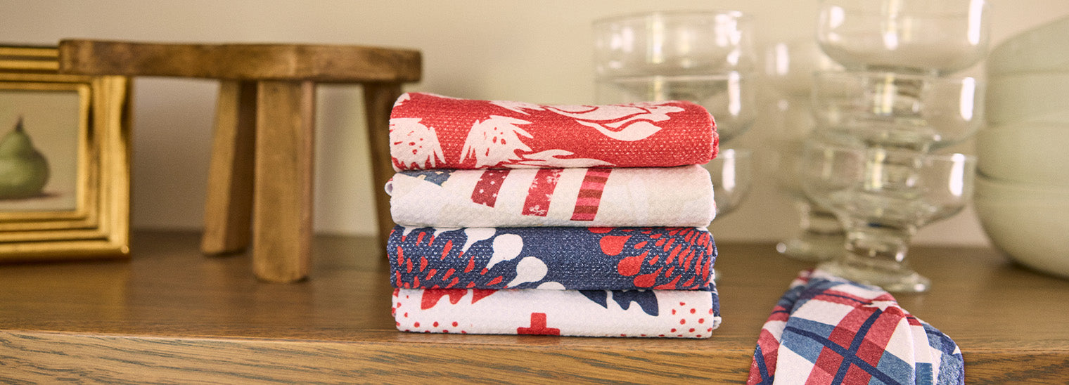 Four folded red, white, and blue patterned cloth napkins stacked on a wooden surface, with glassware, a framed pear painting, and a small wooden stool in the background.