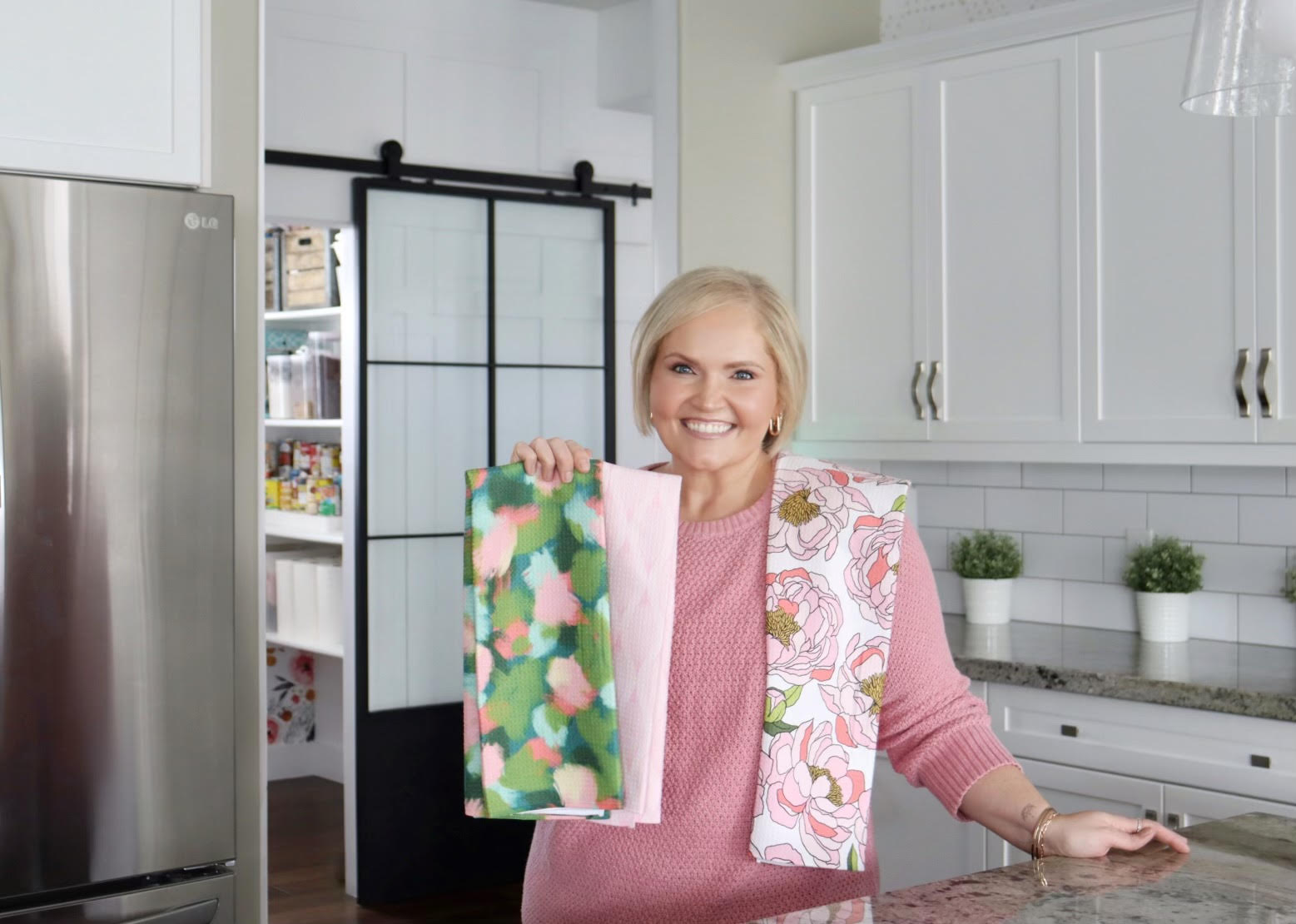 A smiling woman in a pink sweater stands in a bright kitchen, holding up two colorful patterned towels—one with green and pink abstract shapes, the other with pink floral prints.