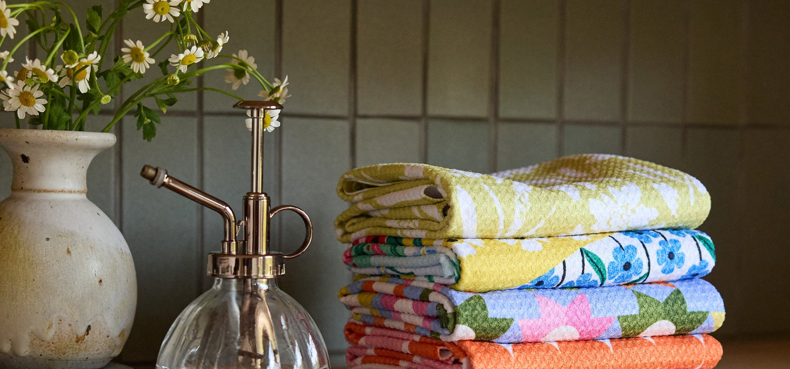 A stack of colorful, patterned towels is neatly folded next to a vintage glass spray bottle and a vase of white daisies on a wooden surface, with a tiled wall in the background.