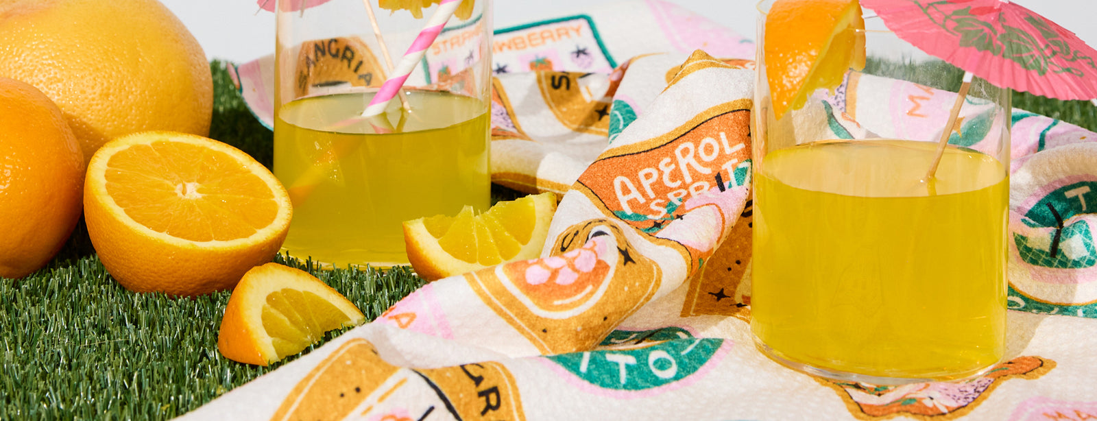 Two glasses of yellow-orange drink with orange slices and umbrellas sit on a decorative cloth, surrounded by whole and sliced oranges on green artificial grass.
