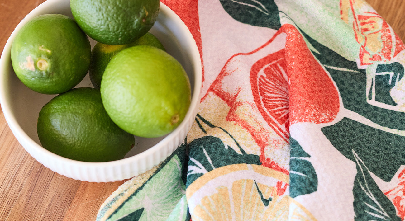 A white bowl filled with green limes sits on a wooden surface next to a colorful, fruit-patterned towel featuring illustrations of lemons and grapefruits.