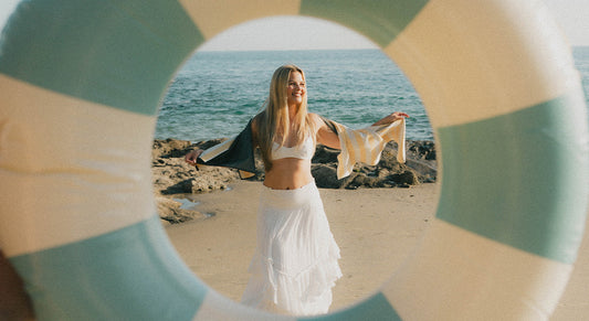 A smiling woman in a white skirt and bikini top stands on a sandy beach, framed through a blue and white lifebuoy, with rocks and the ocean in the background.