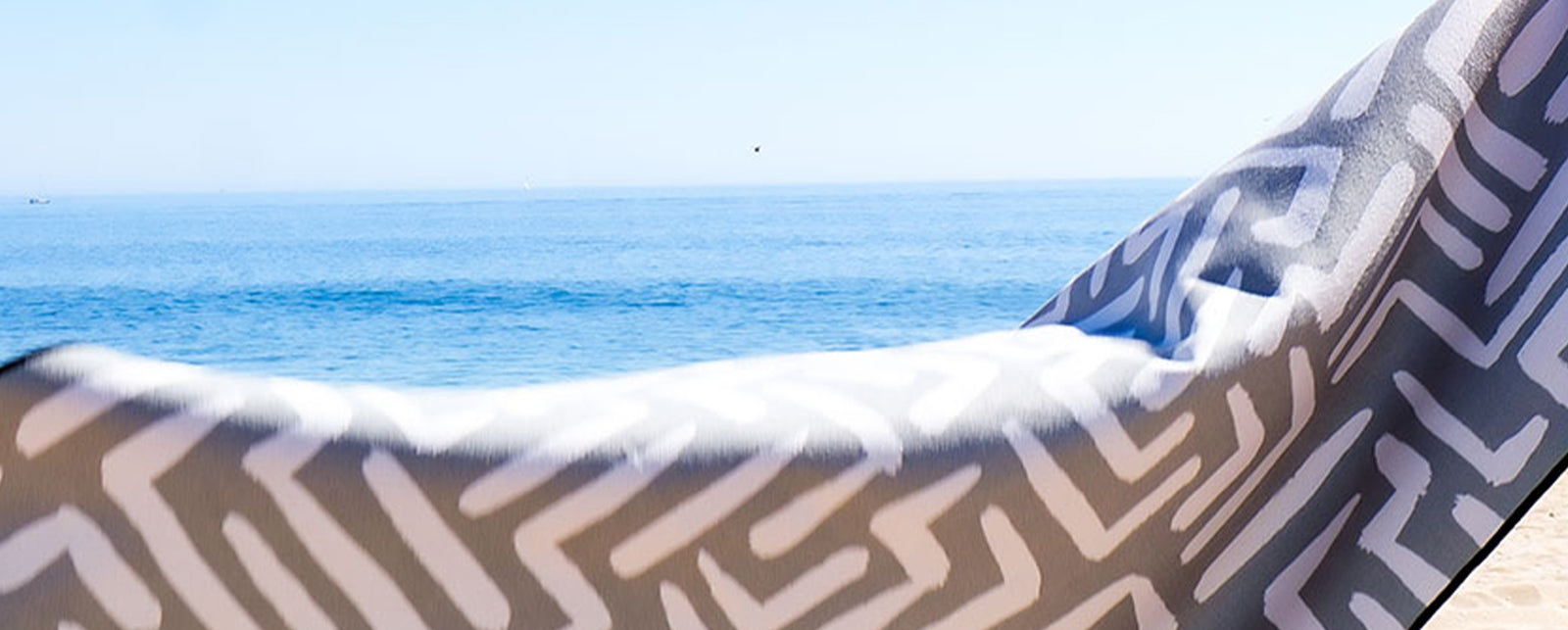 A close-up of a patterned beach towel draped over a chair or hammock with a clear blue ocean and sky in the background.