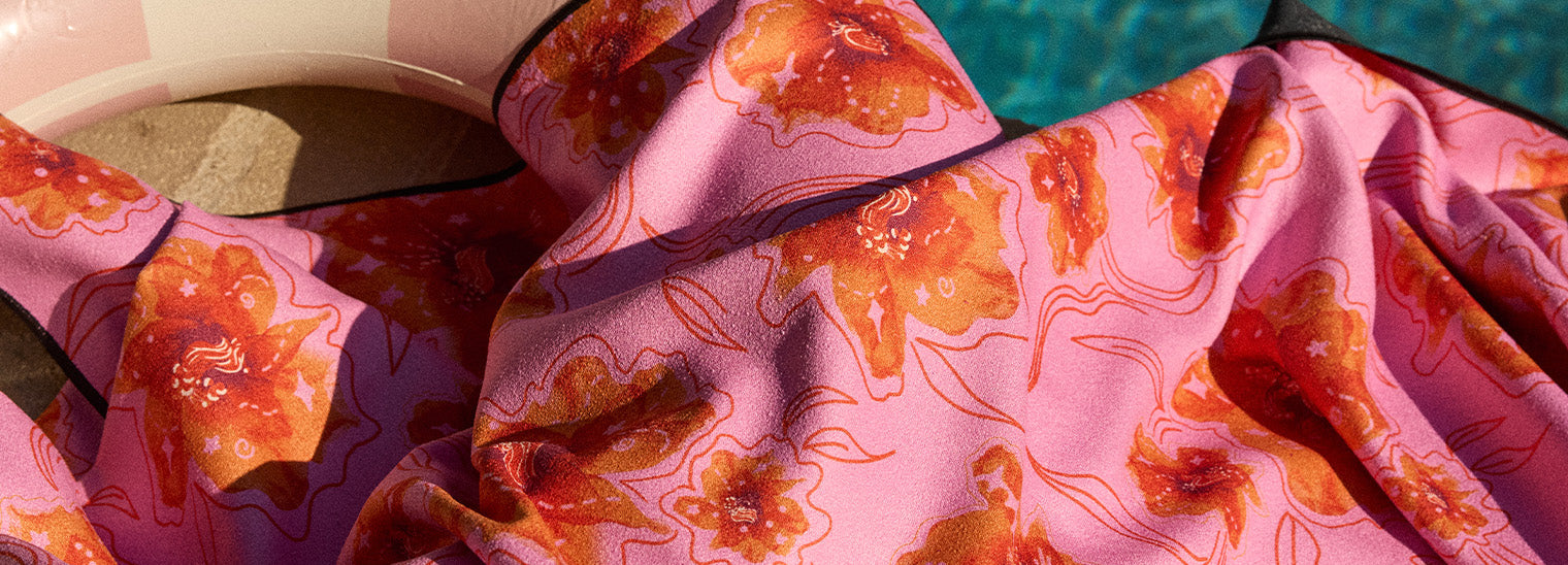 A close-up of a pink and orange floral-patterned fabric draped over a poolside chair with a beige pool float visible in the background near a blue swimming pool.