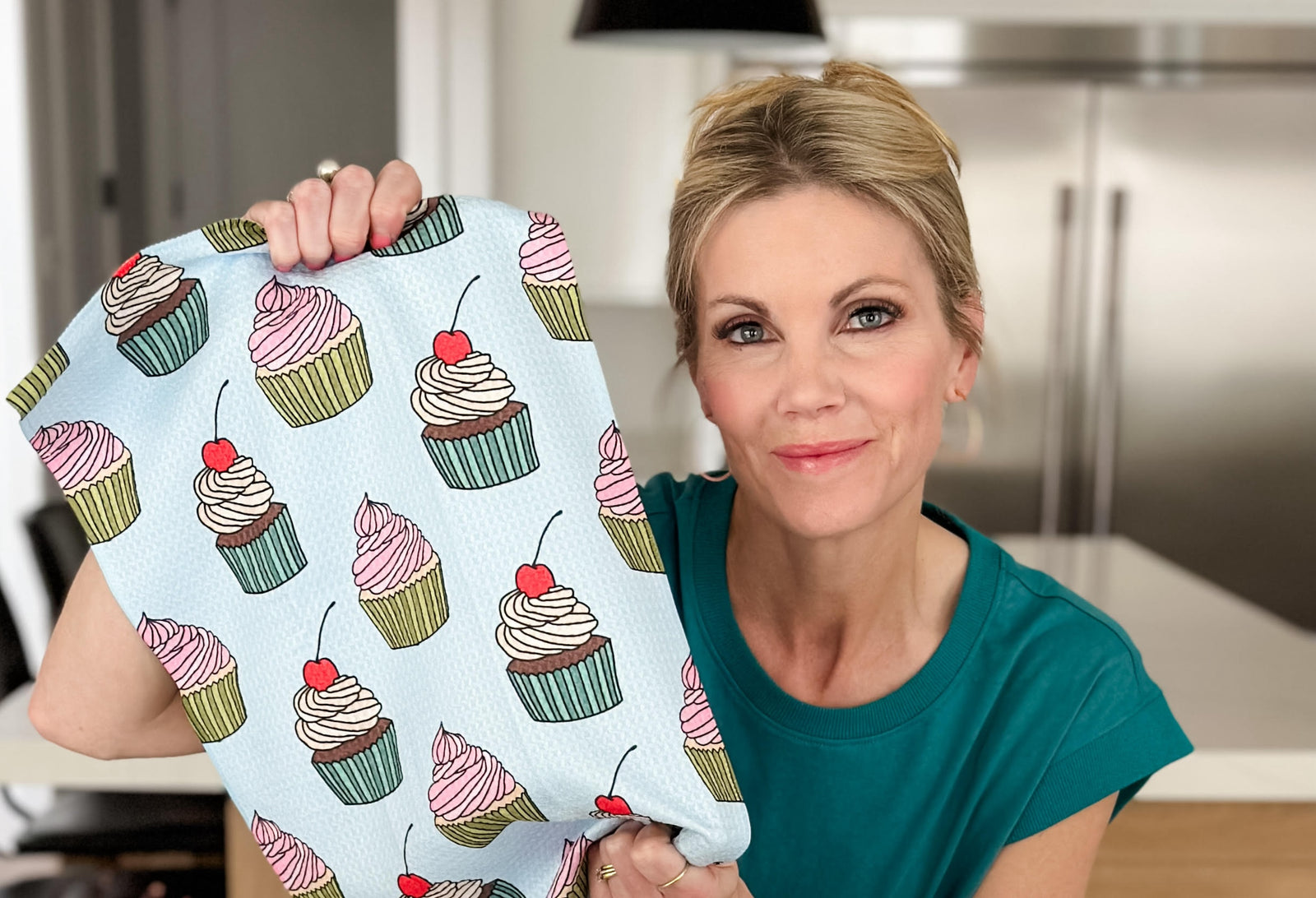 A woman in a teal shirt holds up a light blue towel decorated with colorful cupcakes, standing in a modern kitchen with stainless steel appliances in the background.
