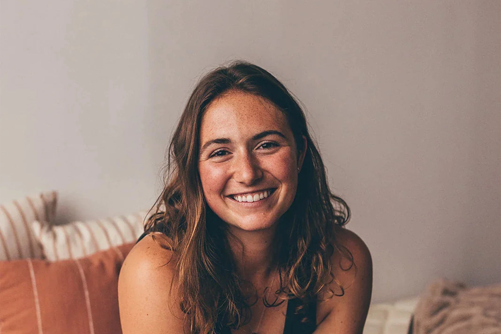 A young woman with long brown hair smiles warmly while sitting indoors on a bed with striped and solid-colored pillows, against a neutral background.
