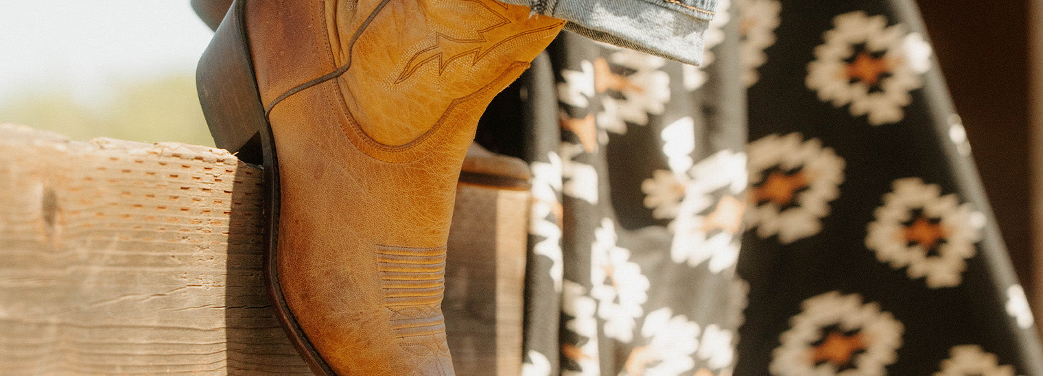 A close-up of a person wearing tan cowboy boots with denim jeans, sitting on a wooden fence next to someone in a black and white patterned dress.