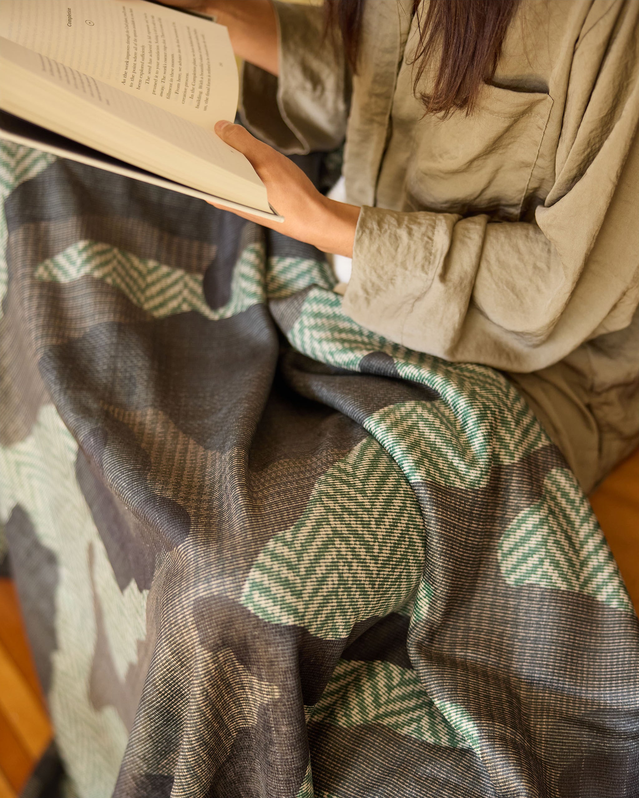 Wrapped in Geometry’s Calloway throw blanket in green, brown, and beige, a person sits reading; only their hands, book, and some hair are visible above the soft drape.
