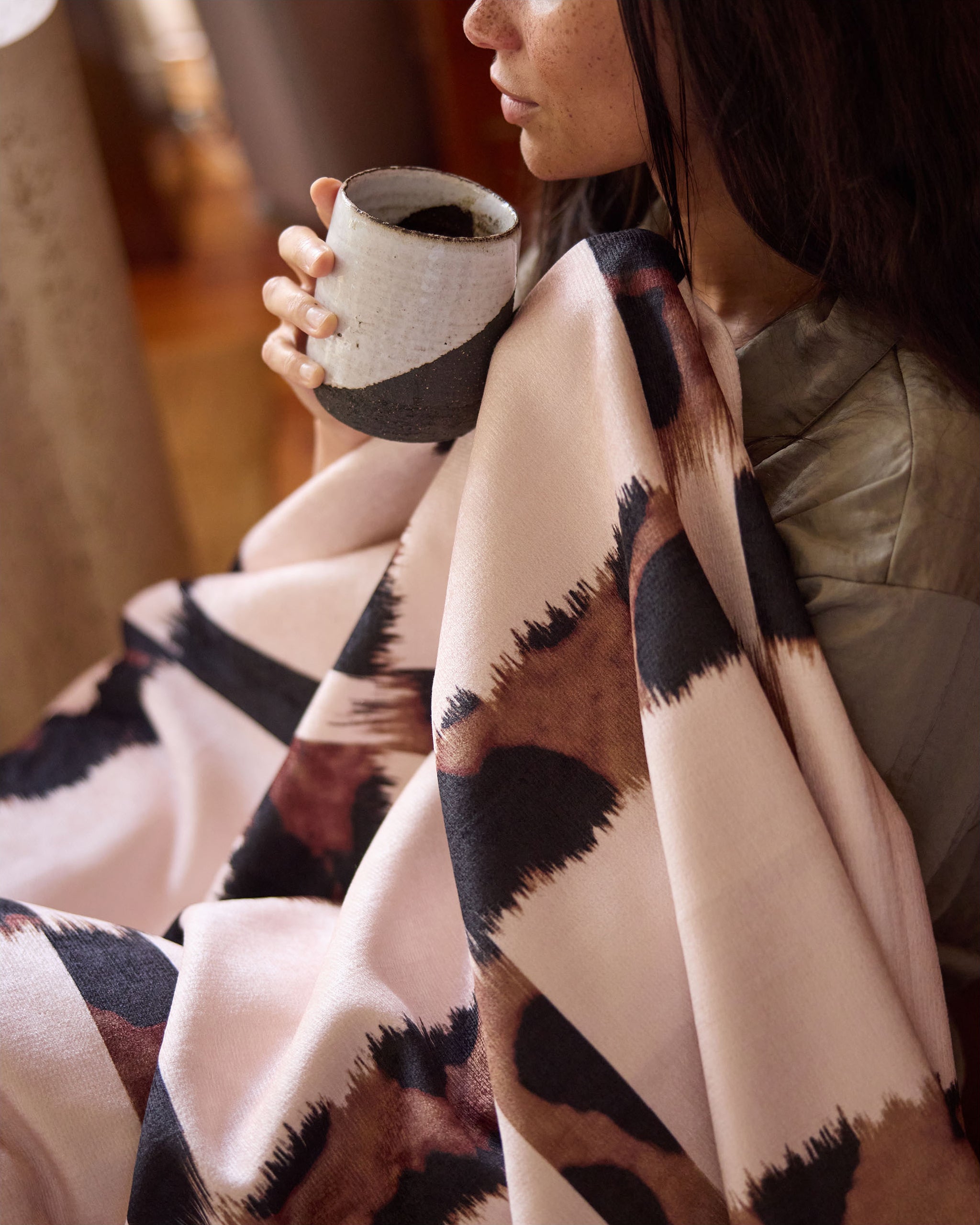 Wrapped in the soft Cheetah Grid throw blanket by Geometry, a person holds a ceramic mug close to their face, creating a cozy, relaxed atmosphere.