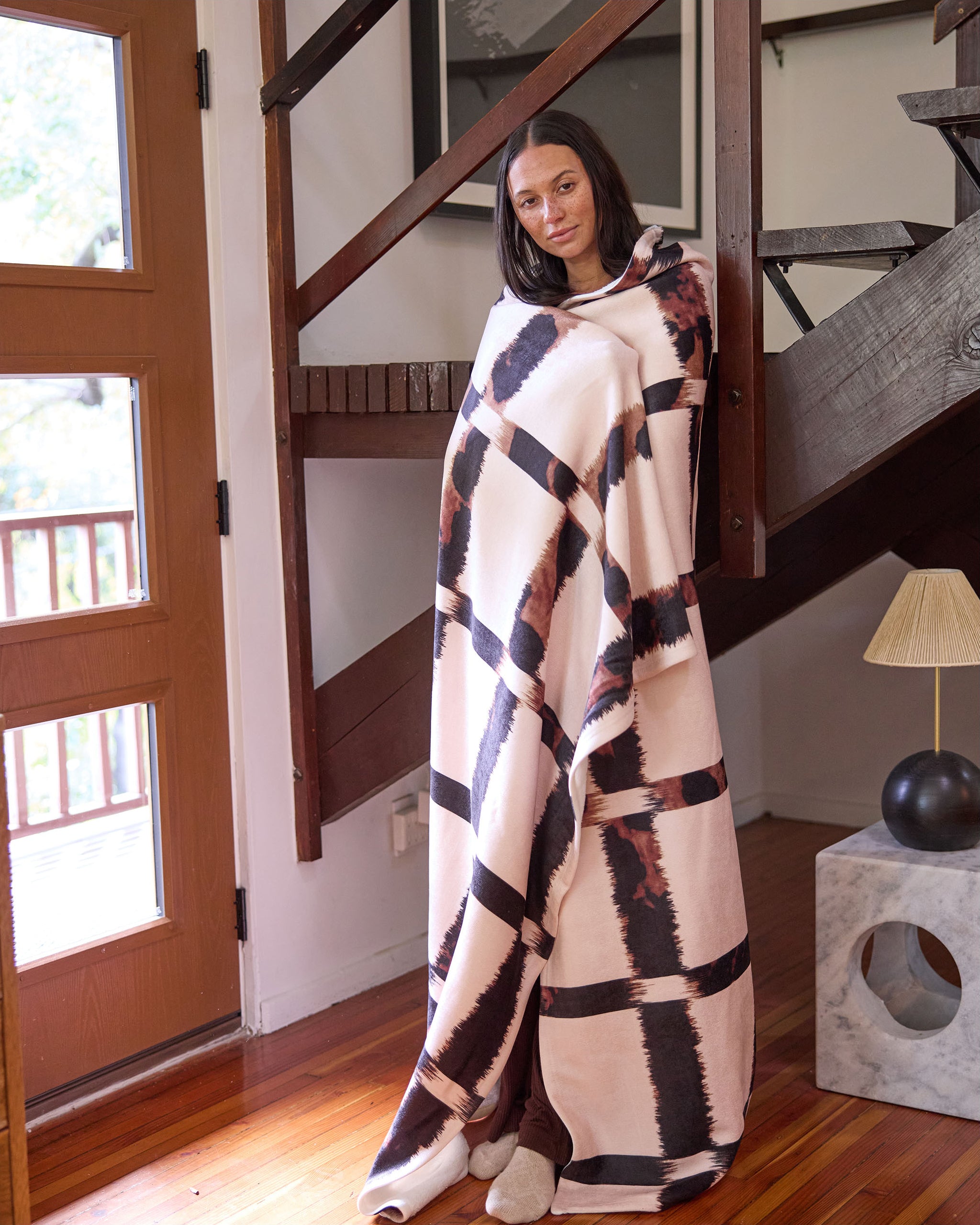 A woman stands indoors wrapped in Geometry’s Cheetah Grid blanket, featuring brown, black, and cream patterns, leaning against a wooden staircase on a hardwood floor in a modern, cozy room.