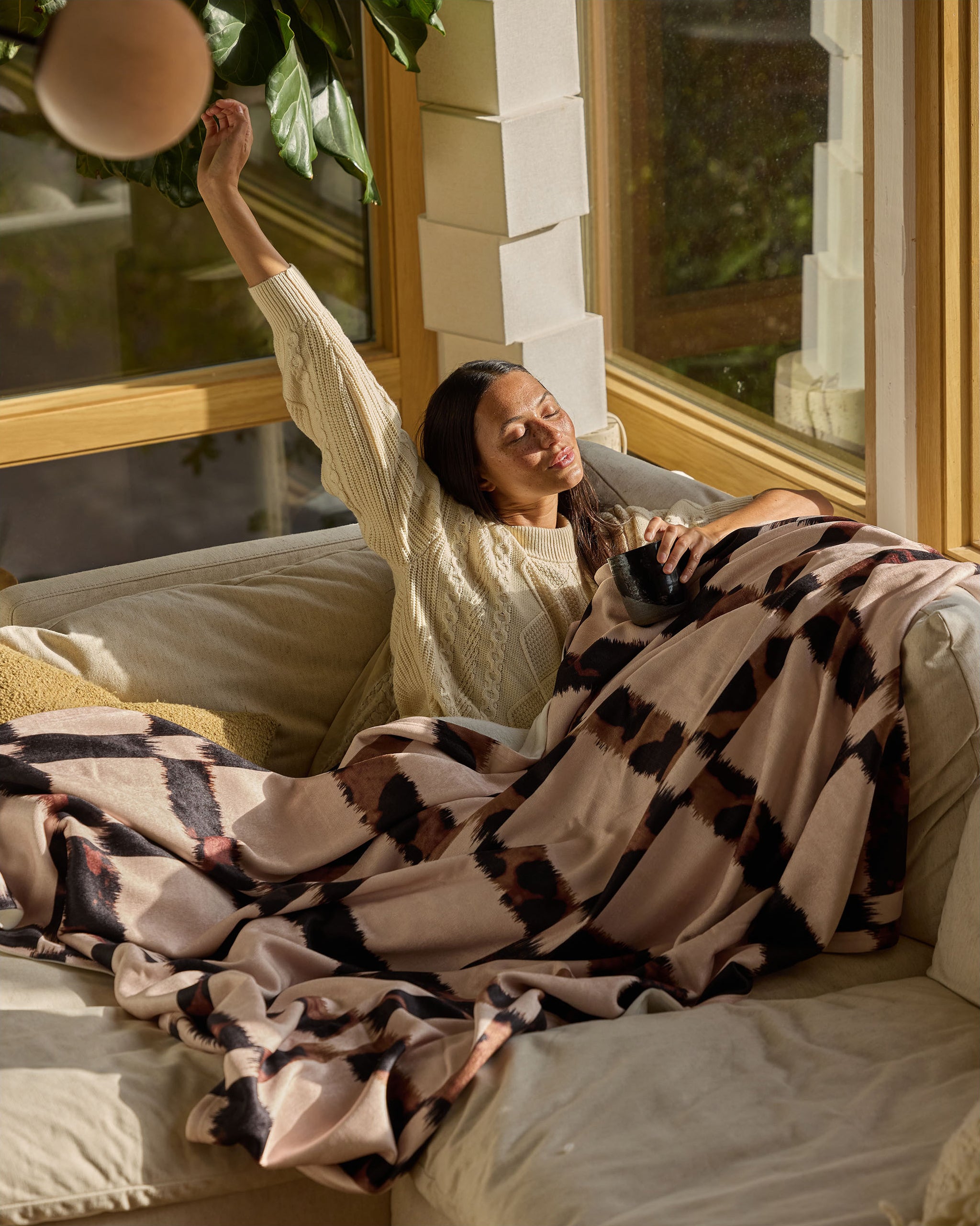 A woman relaxes on a cozy cream sofa by a sunlit window, stretching with a smile while wrapped in the Geometry Cheetah Grid blanket and holding a black mug. Sunlight creates a warm, inviting atmosphere.