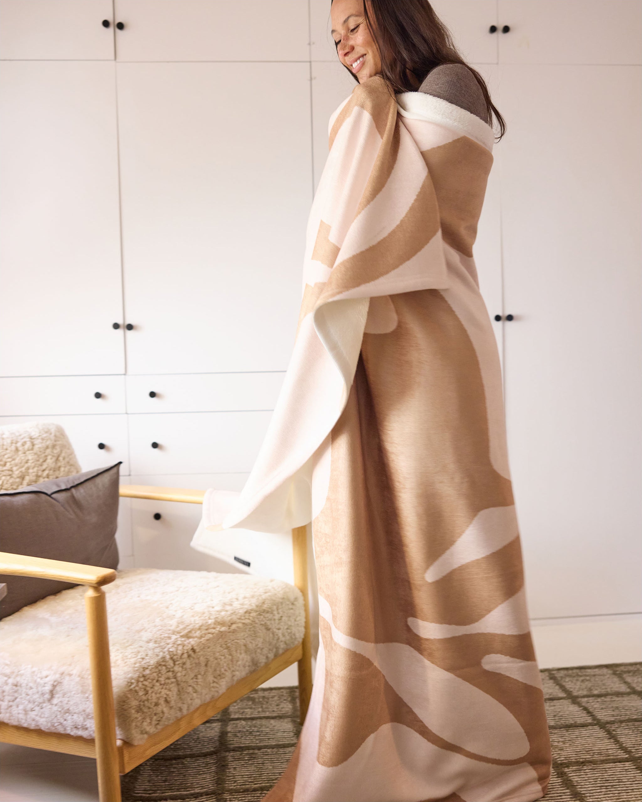 A smiling person stands wrapped in the Cleo travel blanket by Geometry, featuring a beige and white pattern, next to a cushioned wooden chair in a bright room with white cabinets.