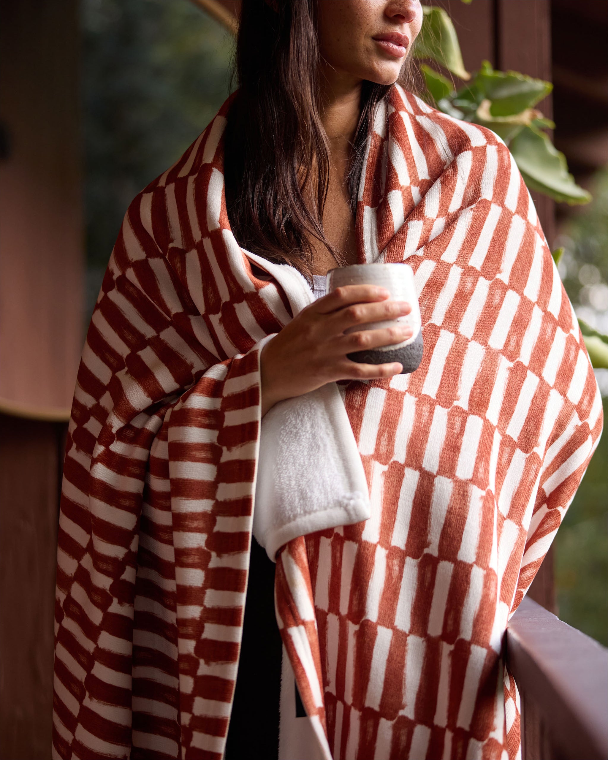 Wrapped in Geometry’s silky soft Clove blanket, a woman stands on a porch holding a mug, lush greenery behind her.