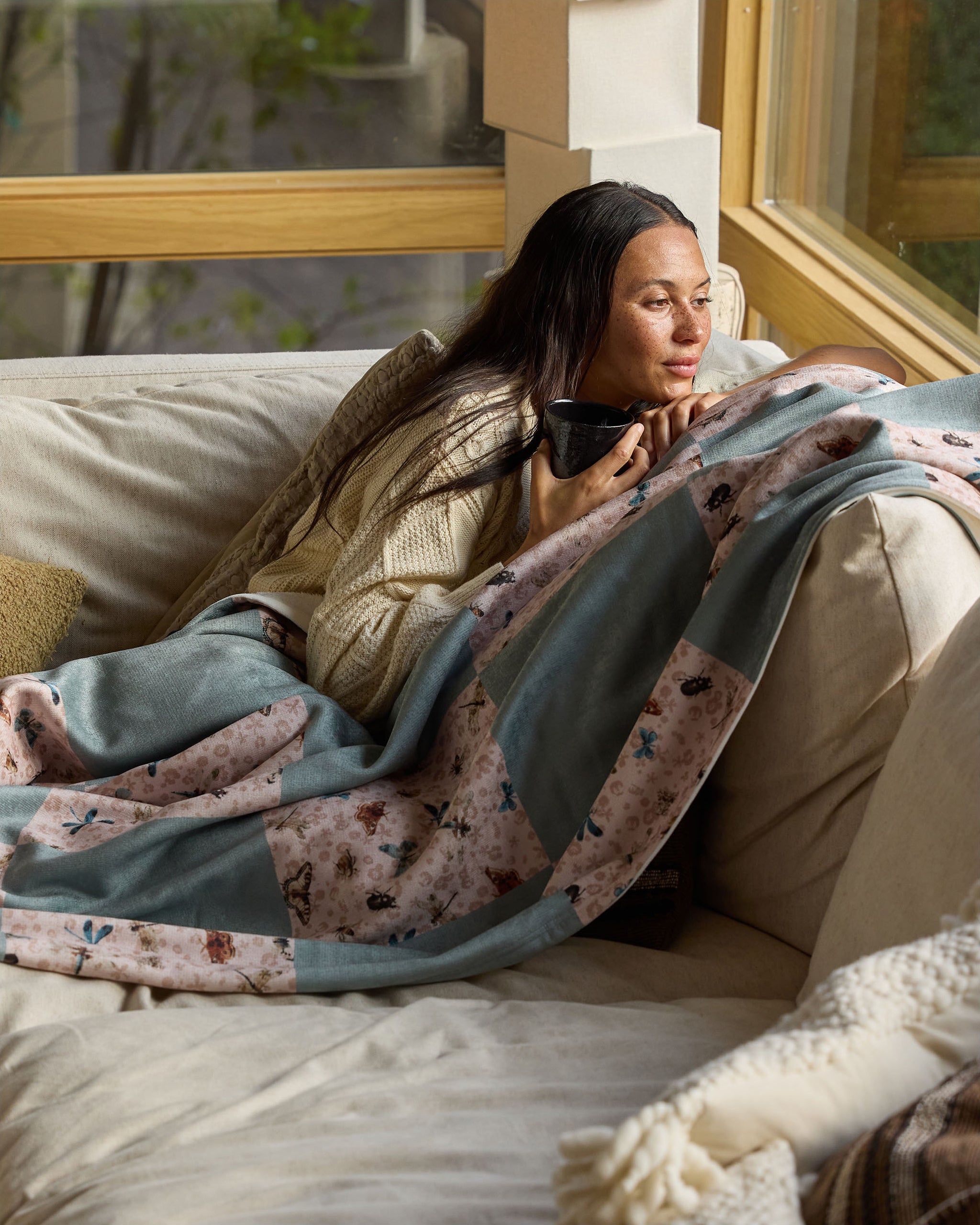 A woman relaxes on a beige couch, wrapped in the soft Critter Check throw blanket by Geometry, holding a mug and gazing out a large window in her warmly lit living room.