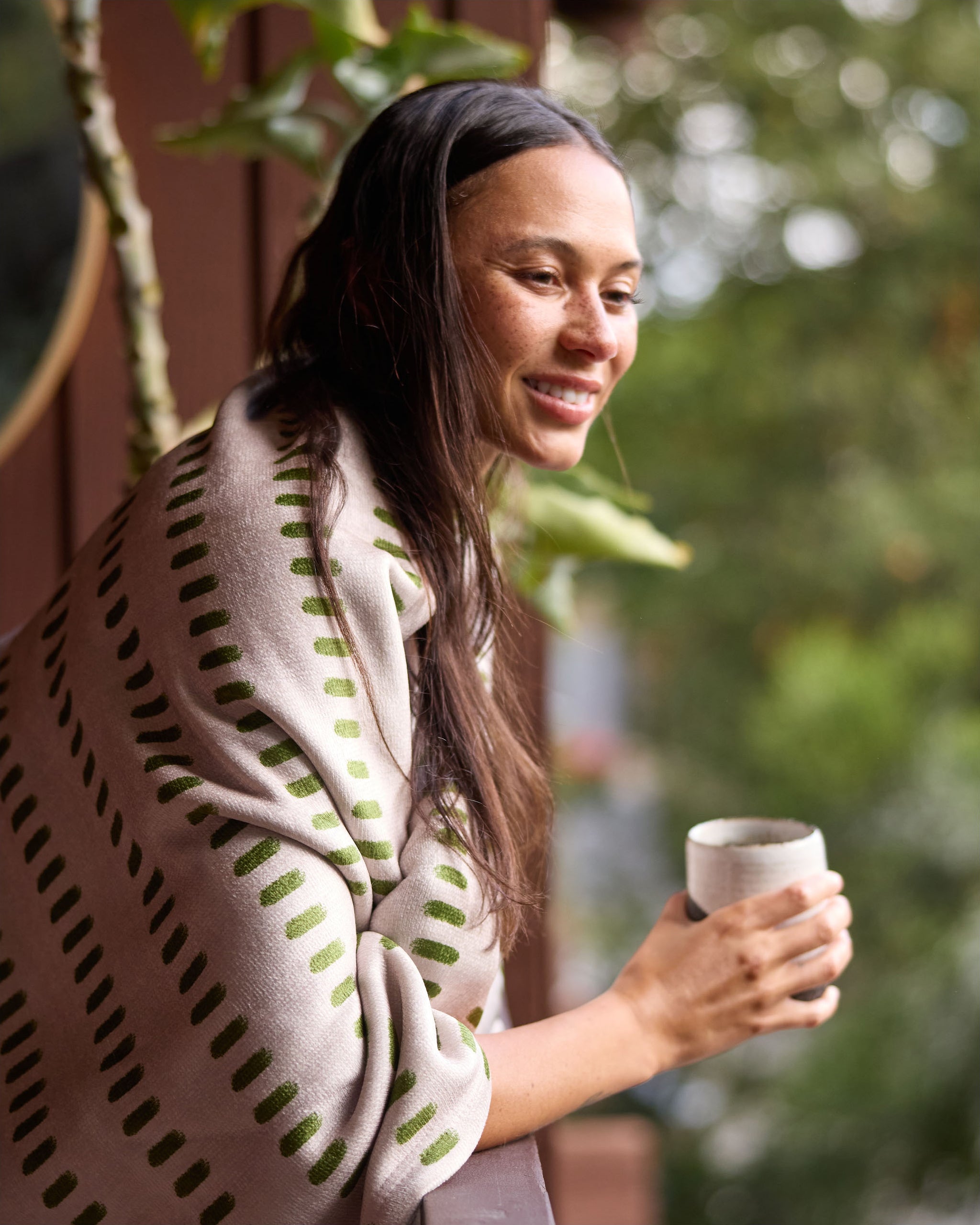 A woman with long dark hair stands on a balcony with greenery, smiling and holding a cup, wrapped in the Gracie recycled blanket by Geometry in a beige and green pattern.