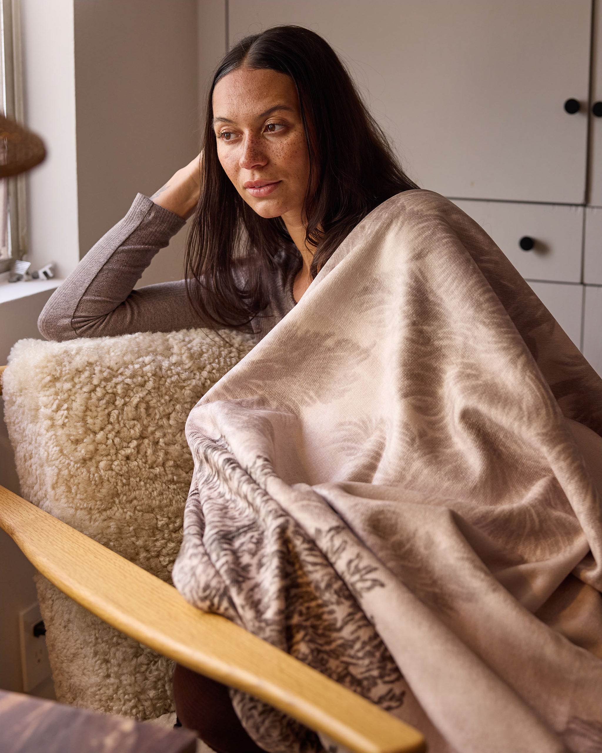 A woman with long dark hair sits in a cozy chair by a window, wrapped in the soft Haven throw blanket from Geometry, looking thoughtfully outside.