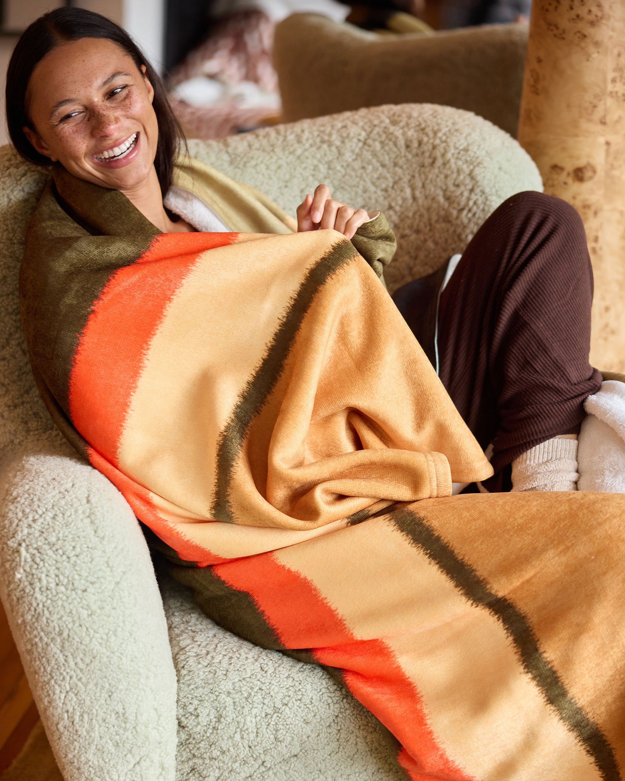 A smiling person sits in a cozy chair, wrapped in Geometry’s Hunting Lodge throw blanket with orange, gold, and green stripes—wearing brown pants and white socks.