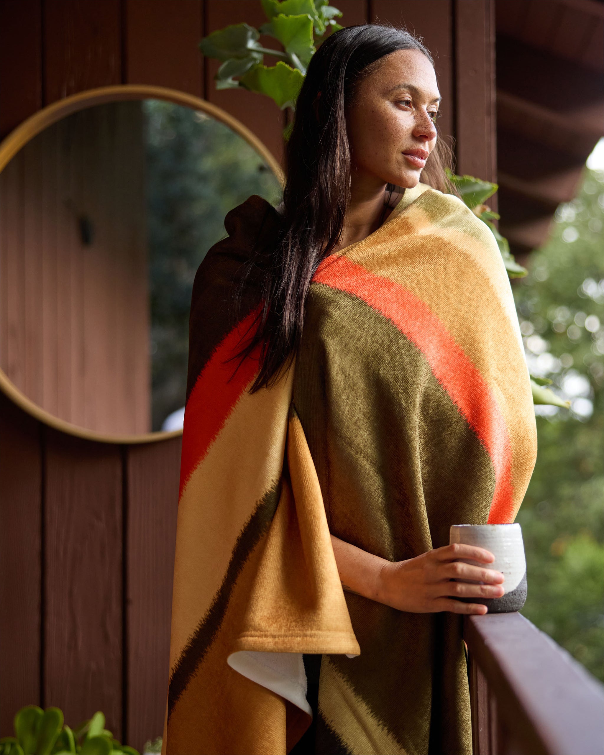 A woman stands on a porch, wrapped in the Geometry Hunting Lodge blanket with earth tones and orange stripes, holding a ceramic mug and gazing thoughtfully. A round mirror and green plants decorate the background.