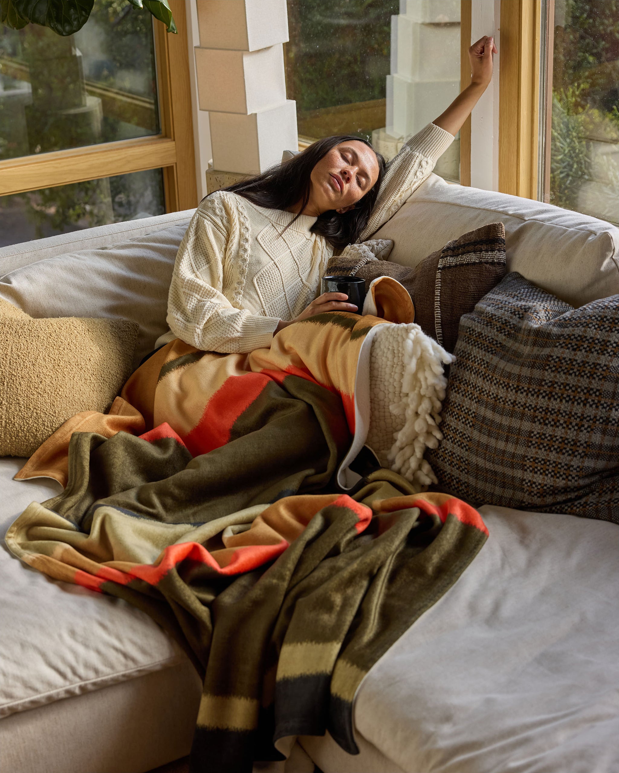 A woman sleeps on a beige couch, wrapped in the Geometry Hunting Lodge throw—a soft blanket with orange, green, and tan stripes. Cozy pillows are behind her as she rests by the window in a cream sweater.