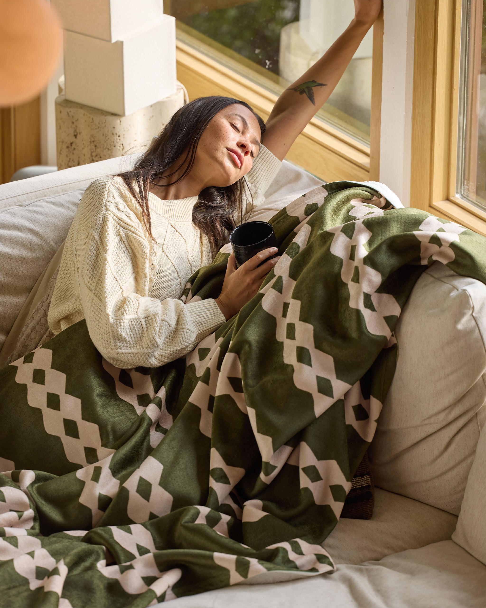 A woman relaxes on a couch, cozy under the Geometry Olive Orchard throw blanket in green and cream patterns, holding a black mug. She wears a cream sweater and rests her arm on the nearby window.