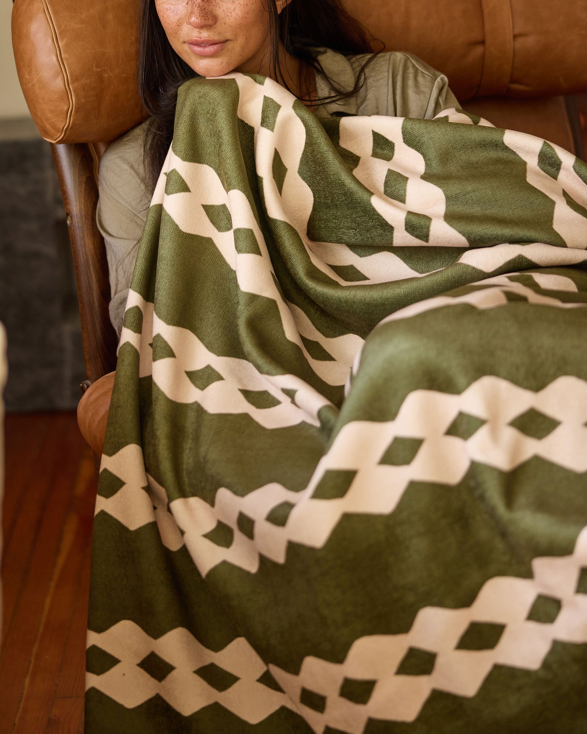 A person sits on a brown leather chair, partially covered by Geometry's Olive Orchard throw blanket, which features a cream geometric pattern of interlocking diamonds and lines. Only the lower half of their face is visible.