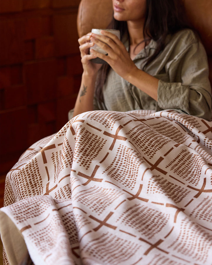 Wrapped in the silky soft Theo blanket by Geometry, a person relaxes in a cozy chair with a mug in both hands. Warm-toned wooden walls add to the inviting, comfortable ambiance.