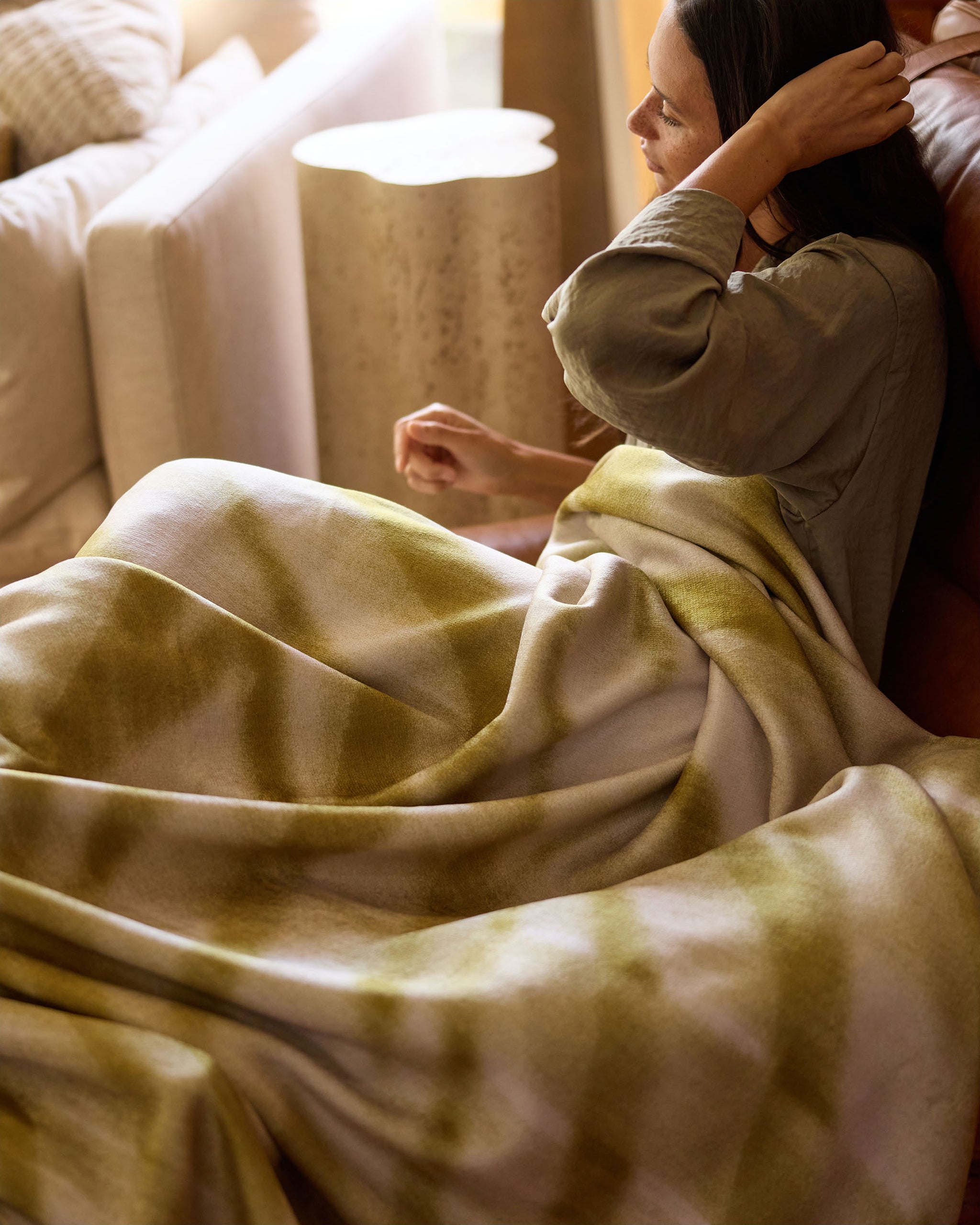 A person sits on a couch, partially covered with the green and white Zain ultra-soft blanket by Geometry. As sunlight streams through the window, they look to the side and adjust their hair.