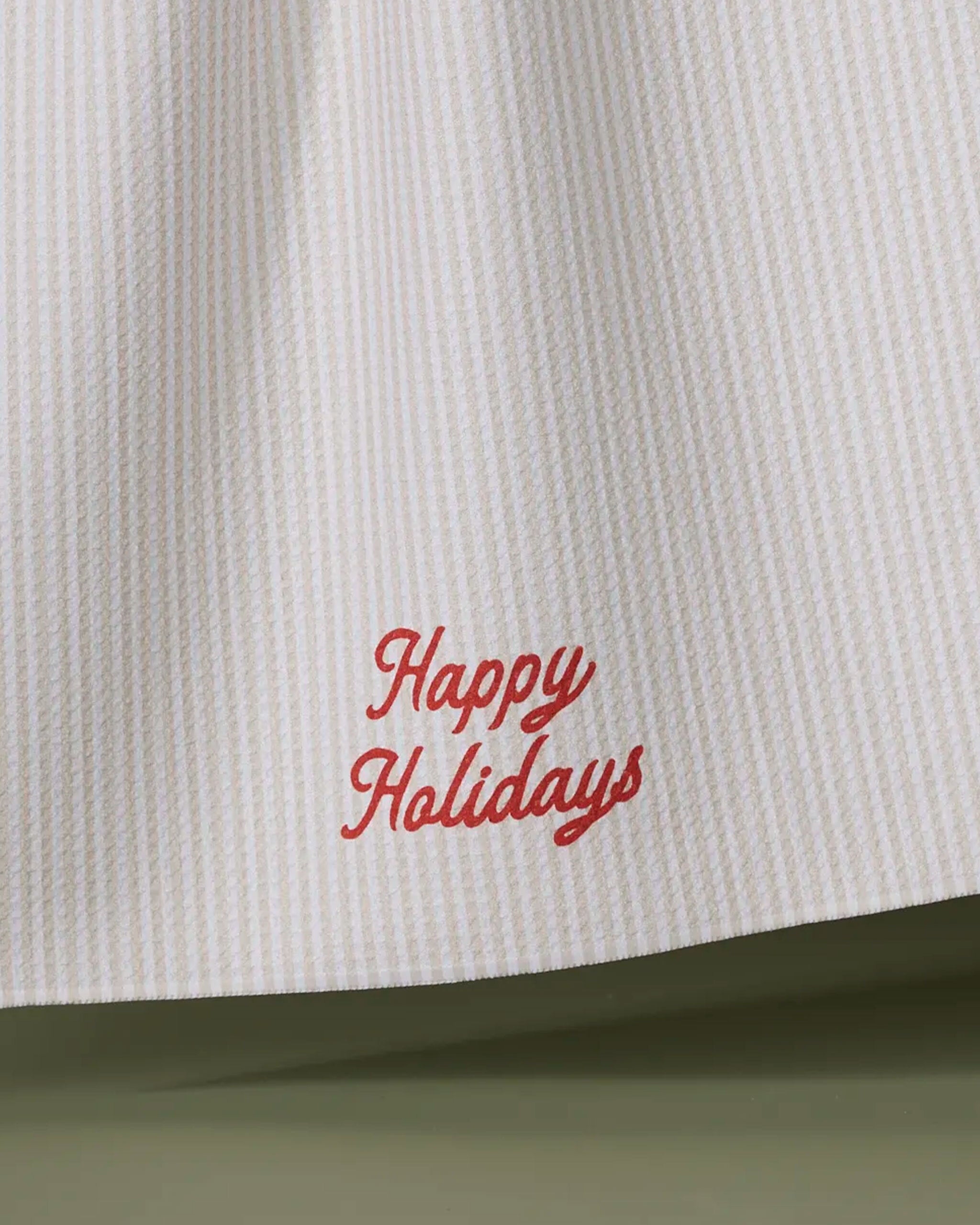 The Geometry Seasons Stripes tea towel is white and textured, made from ultra-absorbent recycled materials, and features "Happy Holidays" embroidered in red cursive near the bottom edge.