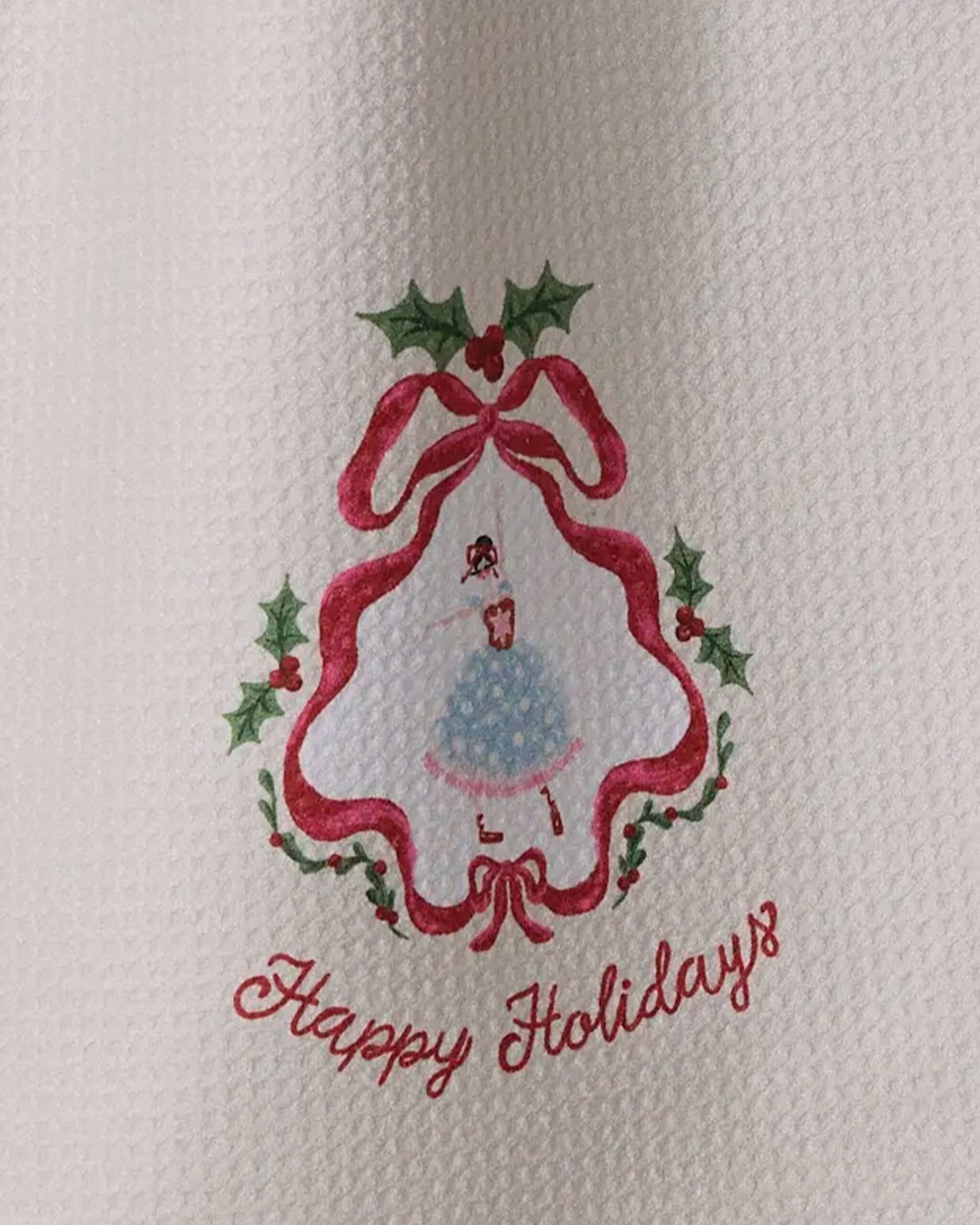 The Geometry "Winter Ballerina" tea towel features a white textured fabric with a Christmas tree outlined in red ribbon and holly, showcasing a girl in a blue dress. "Happy Holidays" is written in red script below.