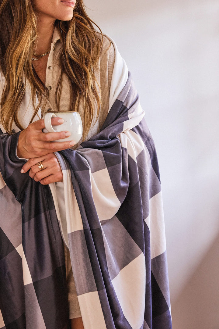 A woman with long hair, wearing a beige button-up, stands against a softly lit background, wrapped in Geometry’s Snowy Gingham blanket made from recycled materials. She holds a white mug and has manicured nails.