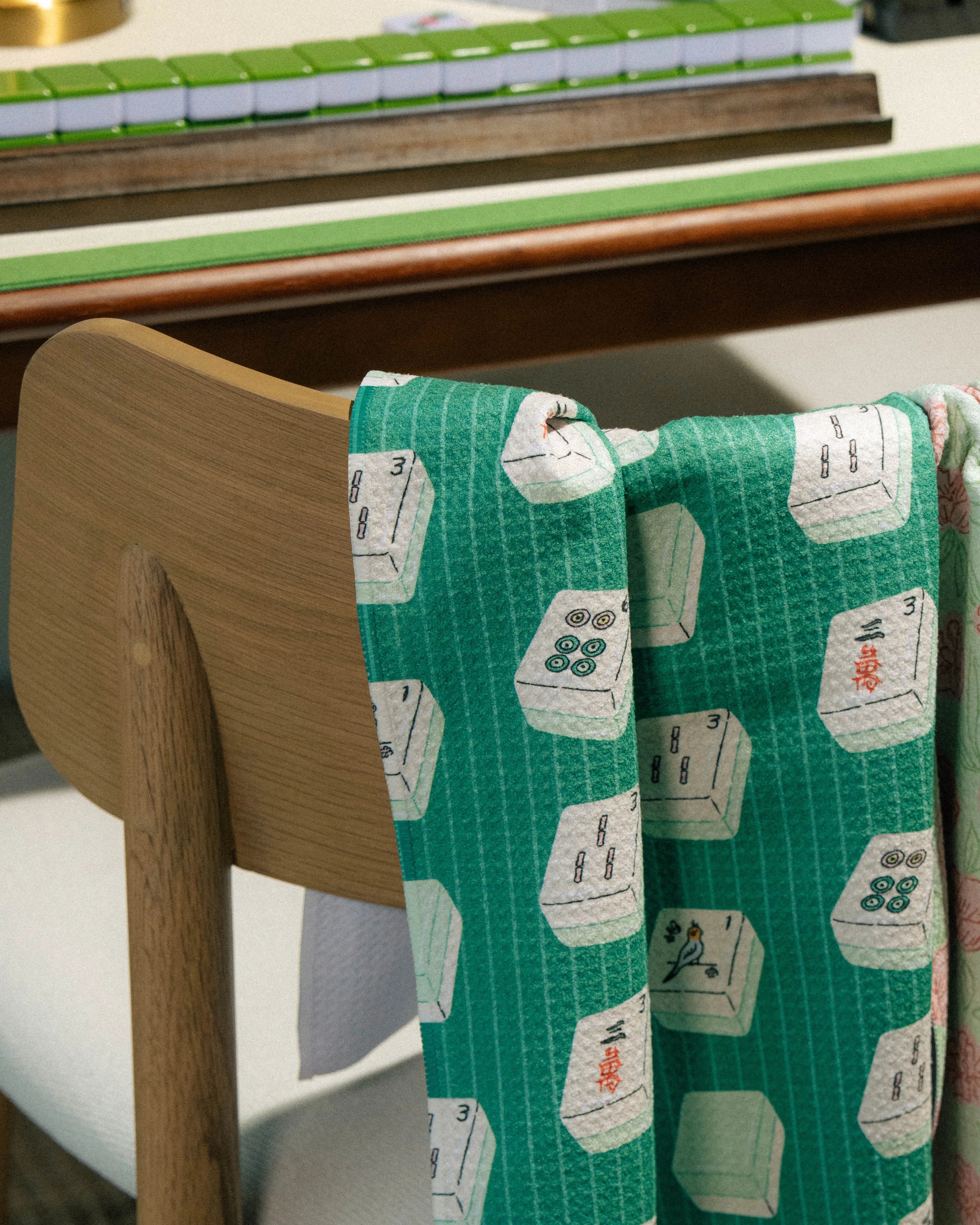 The "Tile Flow" kitchen tea towel by Geometry, showcasing vibrant mahjong tile patterns in green, is draped over a wooden chair with a mahjong set and table in the background. Made from recycled materials, this ultra-absorbent towel blends playful style with eco-friendly functionality.