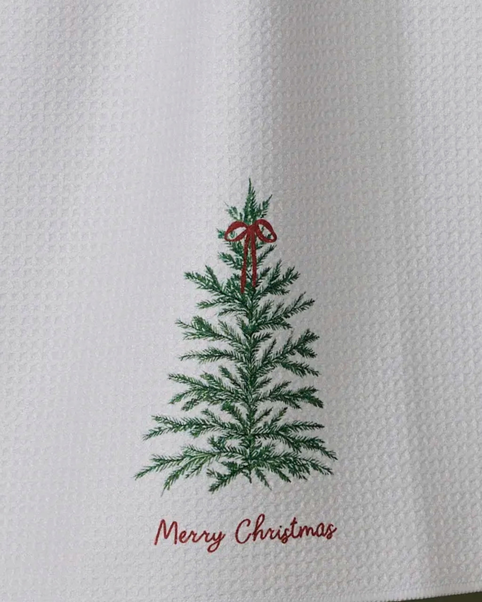The Geometry Redford tea towel features a white textured design with an embroidered green Christmas tree topped by a red bow and "Merry Christmas" written in red script below.