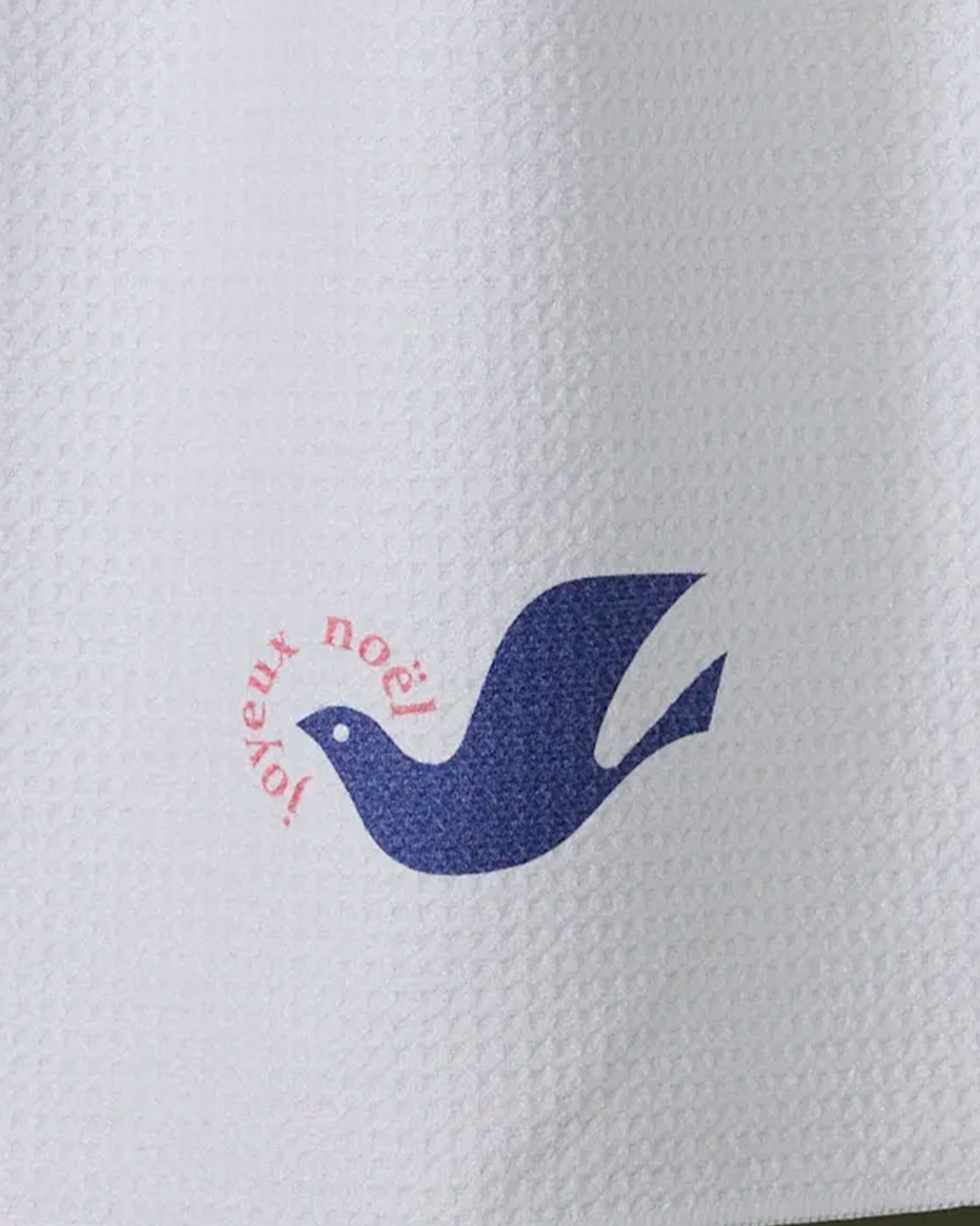 The Geometry Winter Dove kitchen towel is white with a textured design, featuring a simple blue dove illustration and "joyeux noël" in red, arched above the dove.
