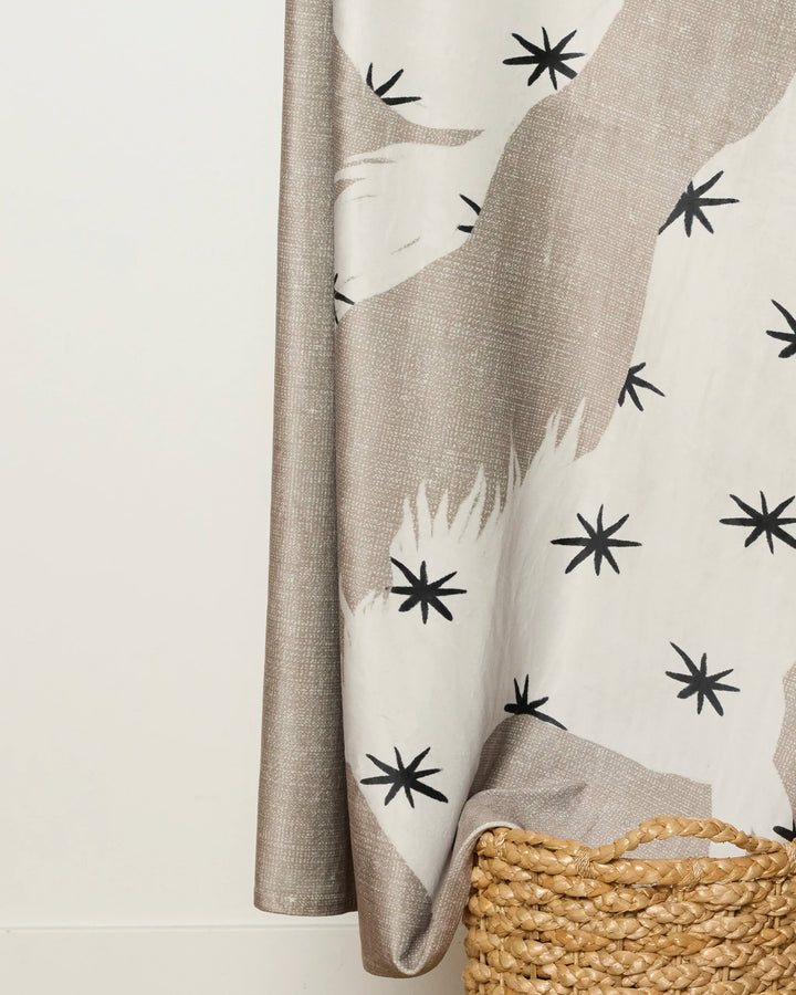 The Unbound curtain by Geometry, featuring beige and white tones with black star patterns, hangs next to a woven basket holding a soft throw on a light floor against a plain white wall.
