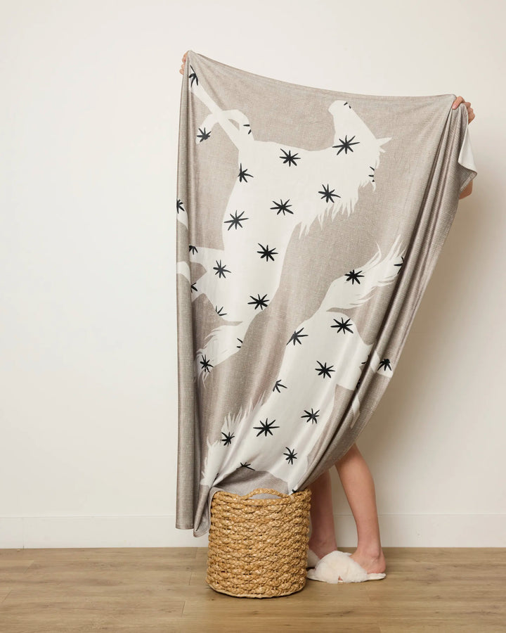 A person stands against a wall, mostly hidden behind the Geometry “Unbound” throw blanket draped over a woven basket. Only their lower legs and slippers are visible.