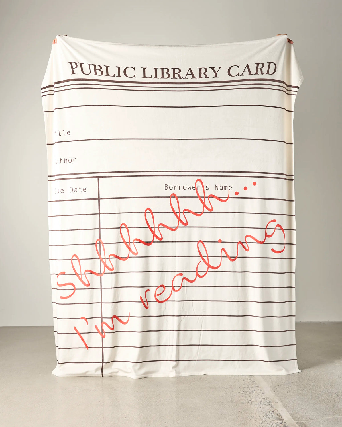 A large, soft Geometry Book Nook throw blanket, printed to look like a public library card and featuring “Shhhhhh... I’m reading” in bold red script, hangs against the wall.