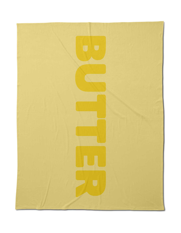 Geometry’s Butter Blanket in light yellow offers silky softness and features “BUTTER” in bold vertical yellow letters. Made from post-consumer recycled materials, it delivers a cozy, eco-friendly touch.