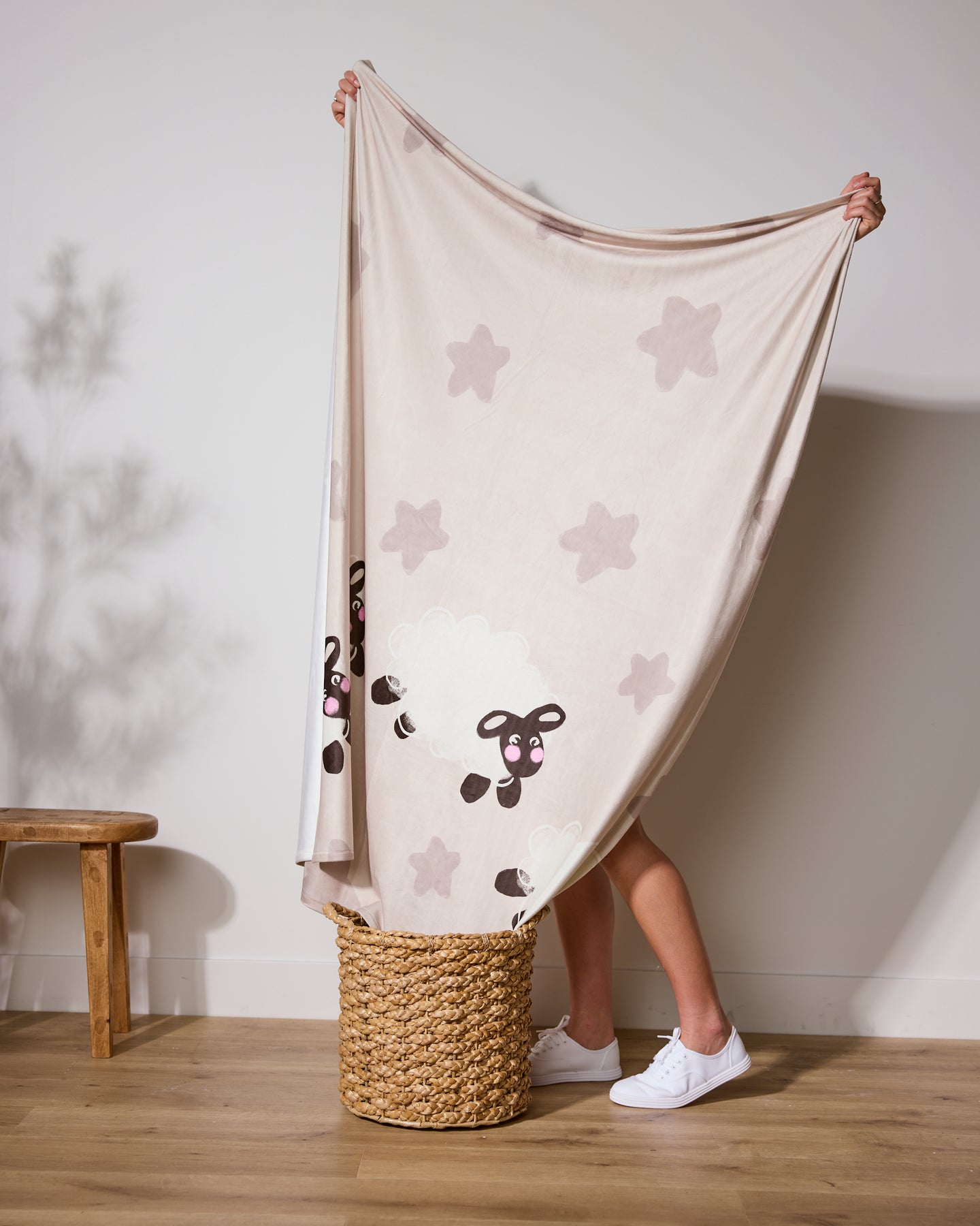 A person stands in a woven basket, holding up Geometry’s Counting Sheep—a soft throw blanket with cartoon sheep and star patterns. Only their legs and white sneakers are visible, with a wooden bench and light wall in the background.