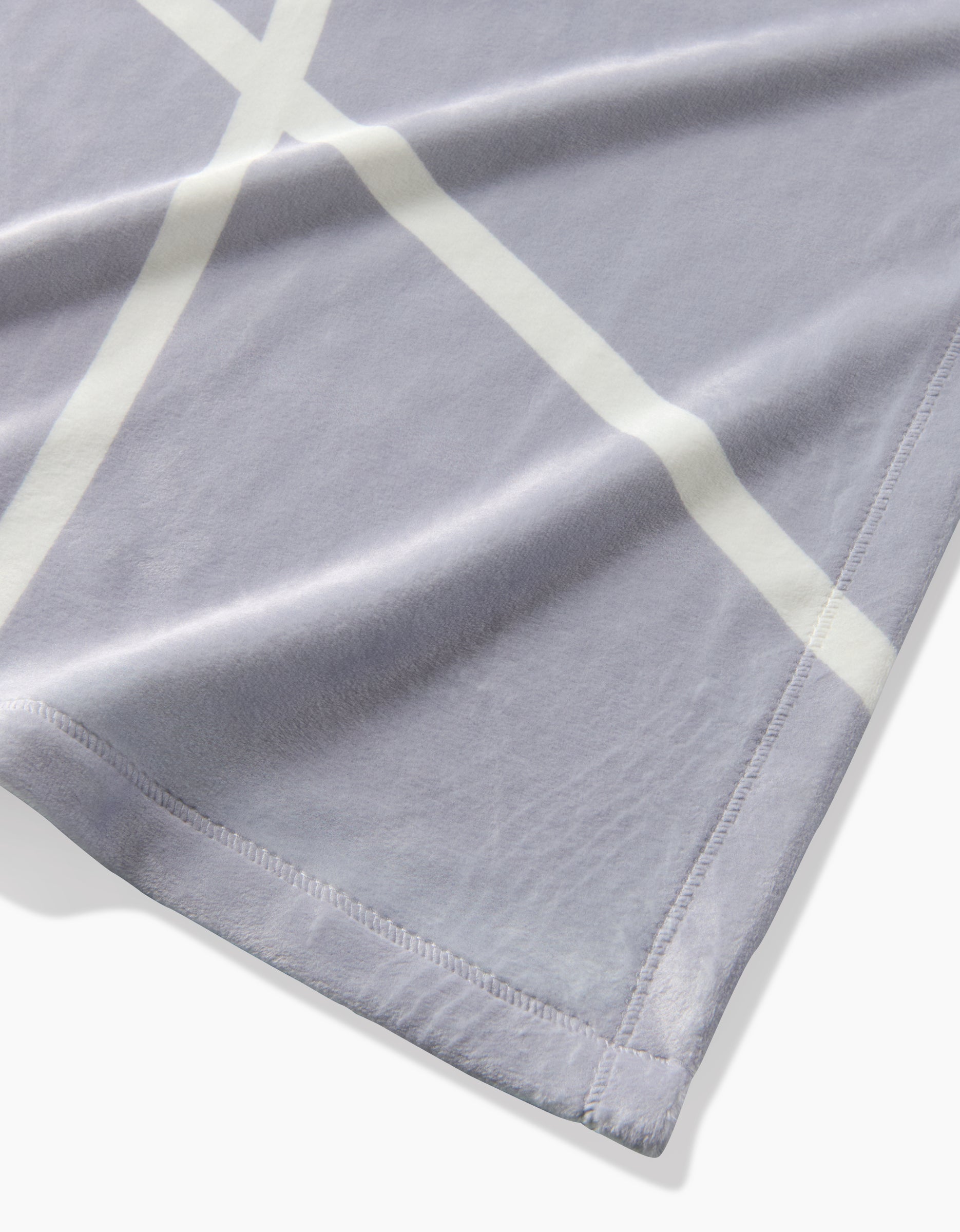 A close-up of Geometrys Crossover blanket, an eco-friendly, soft light gray throw with a white crisscross pattern and neat stitching, shown slightly folded on a white background.